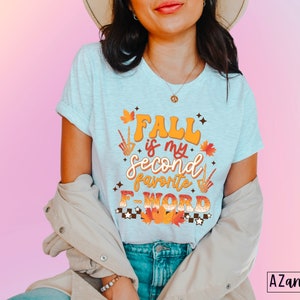 Pu&ograve; includere: T-shirt grigio chiaro con la scritta "Fall is my second favorite F-WORD" in arancione, marrone e bianco. Il design include foglie autunnali e una mano di scheletro. Parte di una collezione a tema autunnale.