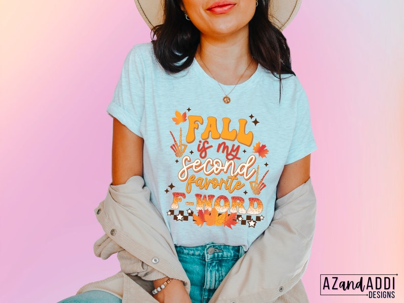 Pu&ograve; includere: T-shirt grigio chiaro con la scritta "Fall is my second favorite F-WORD" in arancione, marrone e bianco. Il design include foglie autunnali e una mano di scheletro. Parte di una collezione a tema autunnale.