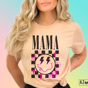 Pu&ograve; includere: Una maglietta color beige con la parola "MAMA" stampata in lettere nere. La maglietta presenta un design a scacchi bianco e nero con un'emoticon sorridente rosa e un grafico a forma di fulmine.