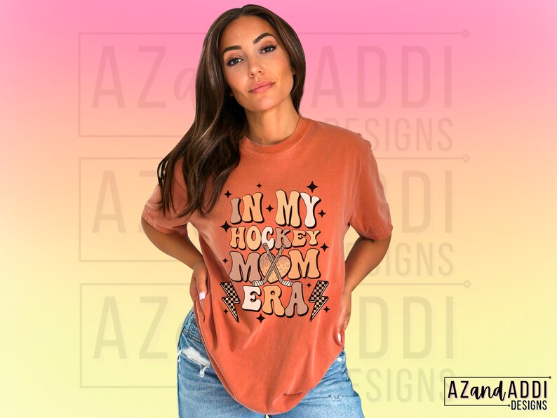 May include: A rust-colored t-shirt with a retro-style graphic that says "In My Hockey Mom Era" with a hockey stick and heart design.