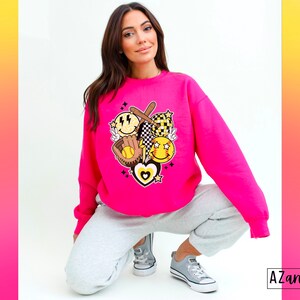 May include: Hot pink sweatshirt featuring a baseball-themed graphic. The graphic includes a baseball bat, glove, smiley faces, and a heart. The sweatshirt is a crew neck style and is paired with gray sweatpants and gray sneakers.