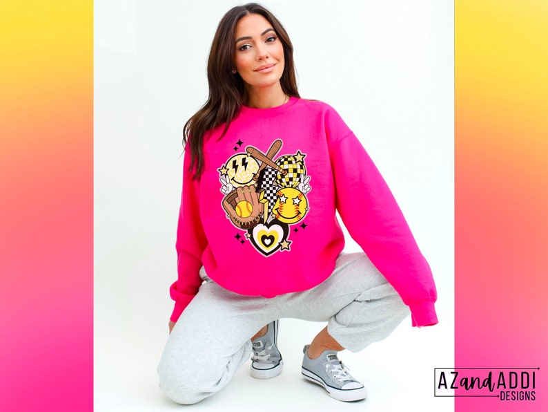 May include: Hot pink sweatshirt featuring a baseball-themed graphic. The graphic includes a baseball bat, glove, smiley faces, and a heart. The sweatshirt is a crew neck style and is paired with gray sweatpants and gray sneakers.