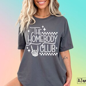 Puede incluir: Camiseta gris con texto blanco que dice "The Homebody Club" con una mano de esqueleto haciendo un signo de rock and roll. El texto est&aacute; rodeado de peque&ntilde;as estrellas blancas y un patr&oacute;n a cuadros.