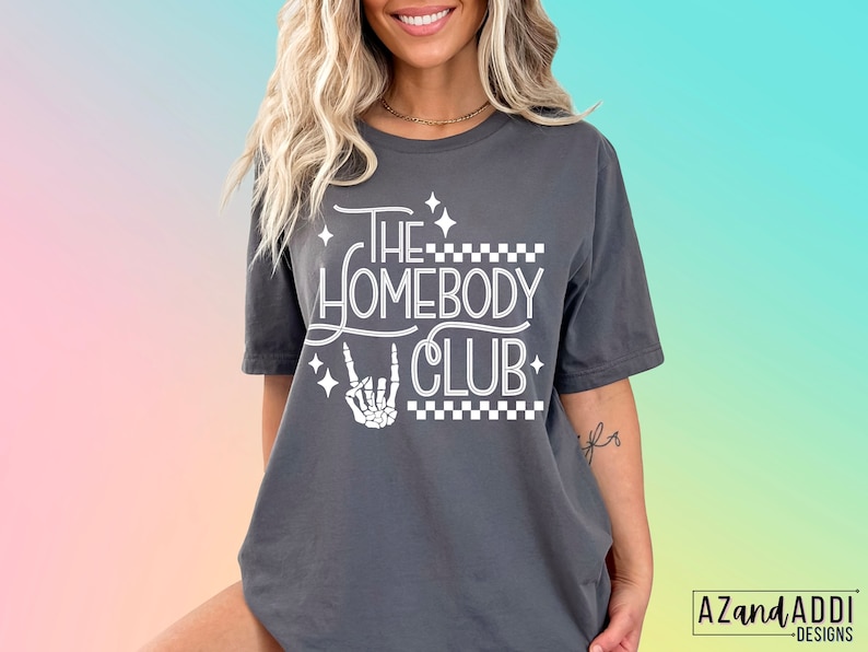 Puede incluir: Camiseta gris con texto blanco que dice "The Homebody Club" con una mano de esqueleto haciendo un signo de rock and roll. El texto est&aacute; rodeado de peque&ntilde;as estrellas blancas y un patr&oacute;n a cuadros.