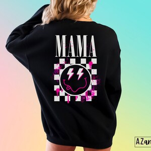 Pu&ograve; includere: Felpa nera con un design a scacchi rosa e bianco, un'emoticon sorridente rosa con fulmini e la parola "MAMA" in lettere bianche.