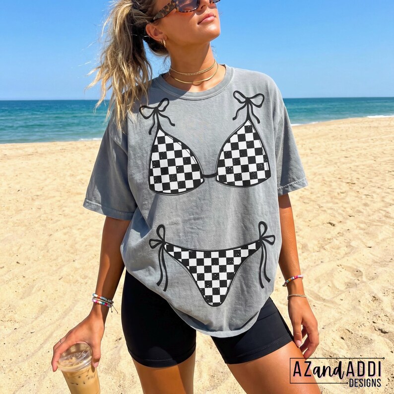 K&ouml;nnte beinhalten: Graues T-Shirt mit einem schwarz-wei&szlig;en karierten Bikini-Design. Das Shirt hat kurze &Auml;rmel und eine lockere Passform. Das Bild ist an einem Strand mit dem Meer im Hintergrund aufgenommen. Der Text "AZandADDI DESIGNS" ist unten rechts sichtbar.