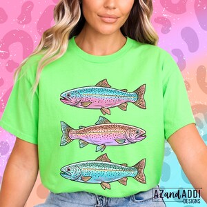 Puede incluir: Una camiseta verde lima con tres ilustraciones de peces coloridos. Cada pez tiene una combinaci&oacute;n de colores &uacute;nica, con tonos de azul, rosa y marr&oacute;n. La camiseta tiene el texto "AZandADDI DESIGNS" en la esquina inferior derecha.