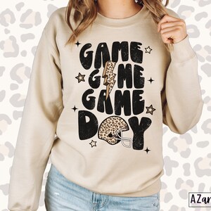 May include: A beige sweatshirt with the text "GAME GAME DAY" in black with a leopard print football helmet and lightning bolt graphic.