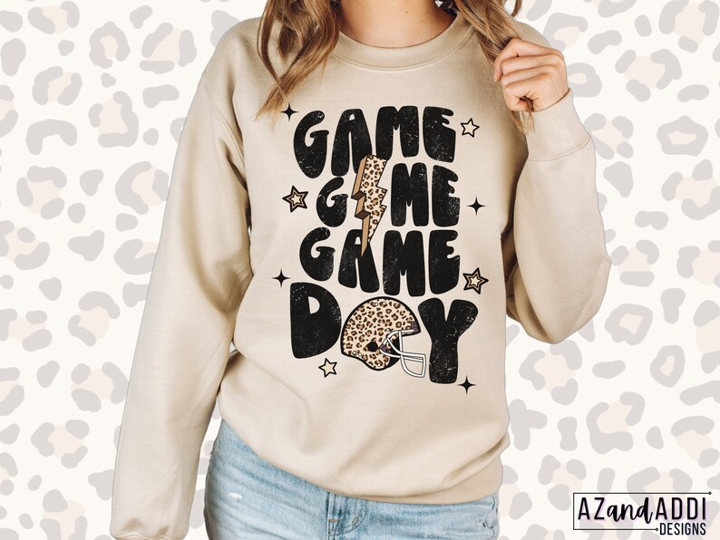 May include: A beige sweatshirt with the text "GAME GAME DAY" in black with a leopard print football helmet and lightning bolt graphic.
