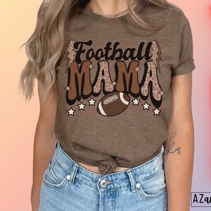 Pode incluir: Uma camiseta marrom com o texto "Football Mama" em uma fonte retr&ocirc; com detalhes de estampa de leopardo e um gr&aacute;fico de futebol. A camiseta est&aacute; sendo usada por uma pessoa com cabelo castanho longo e jeans azul.