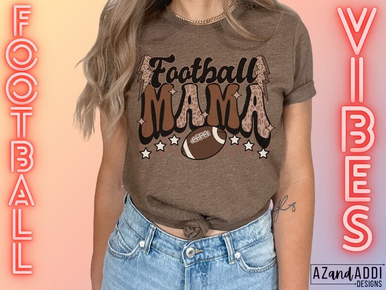 Pode incluir: Uma camiseta marrom com o texto "Football Mama" em uma fonte retr&ocirc; com detalhes de estampa de leopardo e um gr&aacute;fico de futebol. A camiseta est&aacute; sendo usada por uma pessoa com cabelo castanho longo e jeans azul.