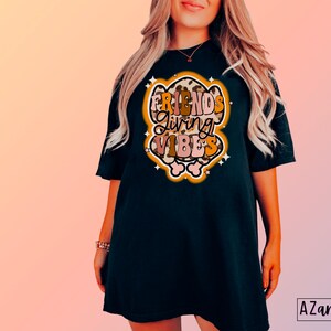 May include: Black t-shirt with the text "FRIENDS GIVING VIBES" in a retro design. The graphic features a leopard print border and star accents. The shirt is a comfortable, oversized fit.