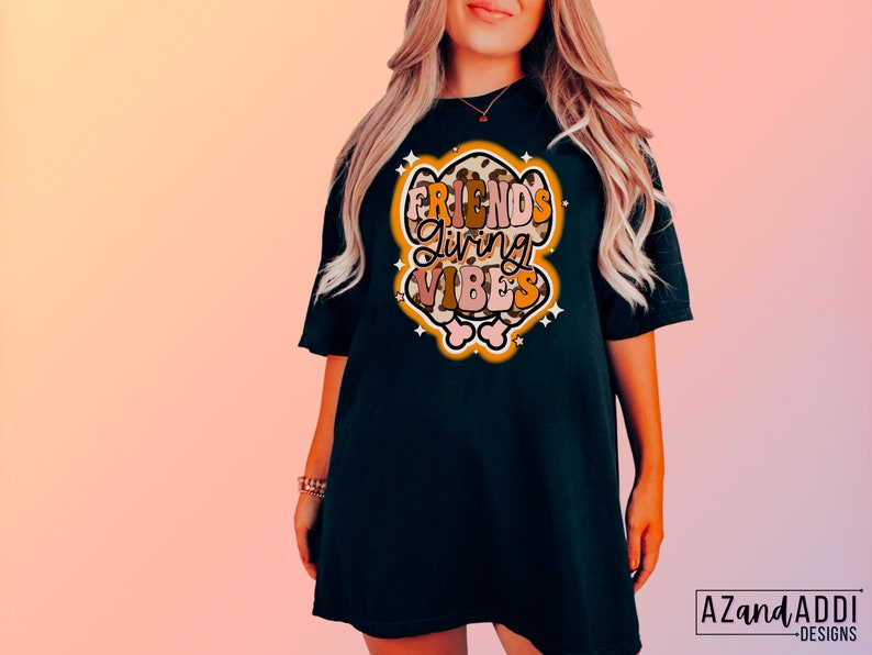 May include: Black t-shirt with the text "FRIENDS GIVING VIBES" in a retro design. The graphic features a leopard print border and star accents. The shirt is a comfortable, oversized fit.