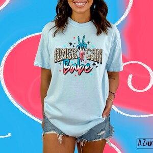 May include: A light blue t-shirt with a graphic print of a peace sign with red, white and blue accents. The text "American Babe" is printed on the shirt in a glittery font.