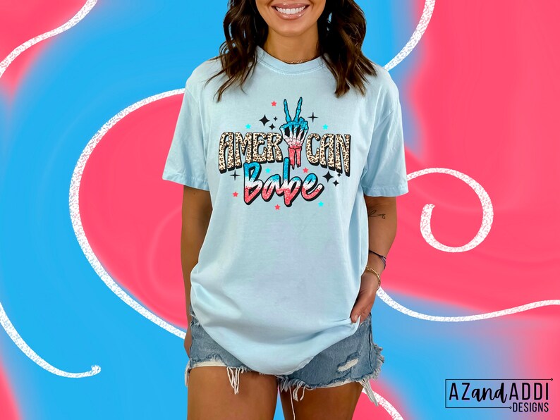 May include: A light blue t-shirt with a graphic print of a peace sign with red, white and blue accents. The text "American Babe" is printed on the shirt in a glittery font.