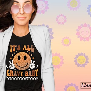 May include: A black t-shirt with a graphic of a smiling face with a leopard print design. The text "It's All Gravy Baby" is printed below the face. The shirt is being worn by a person with long brown hair and pink sunglasses.