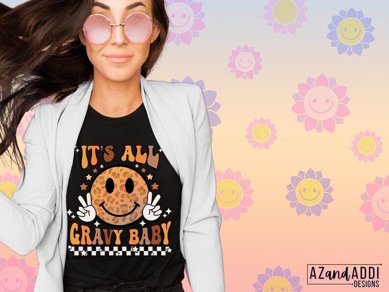May include: A black t-shirt with a graphic of a smiling face with a leopard print design. The text "It's All Gravy Baby" is printed below the face. The shirt is being worn by a person with long brown hair and pink sunglasses.