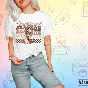 May include: White t-shirt with a brown, black, and white graphic that says "Touchdown Season" with a lightning bolt and footballs. The graphic has a checkered pattern and a leopard print.