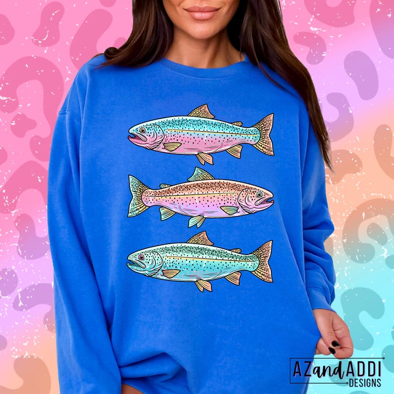 Puede incluir: Sudadera azul con tres ilustraciones de peces coloridos. Los peces tienen una variedad de colores, incluyendo rosa, azul y dorado. La sudadera tiene el texto "AZandADDI DESIGNS" en la esquina inferior derecha.