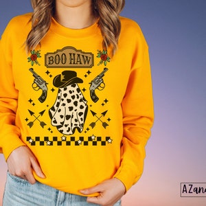 Puede incluir: Sudadera amarilla con un fantasma que lleva un sombrero de vaquero y sostiene dos pistolas. El texto "BOO HAW" est&aacute; en un cartel sobre el fantasma. La sudadera tiene un patr&oacute;n de cuadros en la parte inferior.