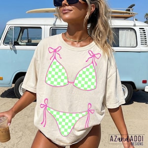 K&ouml;nnte beinhalten: Cremefarbenes T-Shirt mit einem Bikini-Motiv. Das Bikini-Design hat ein gr&uuml;n-wei&szlig;es Karomuster mit rosa Akzenten. Der Text "AZandADDI DESIGNS" ist sichtbar. Ein Surfbrett befindet sich auf einem blauen Van im Hintergrund.