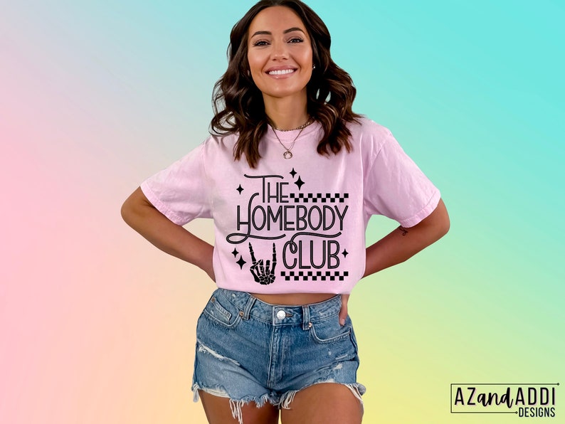 Puede incluir: Una camiseta rosa con texto negro que dice "The Homebody Club" con una mano de esqueleto haciendo un signo de rock and roll. La camiseta tiene un patr&oacute;n a cuadros y estrellas.