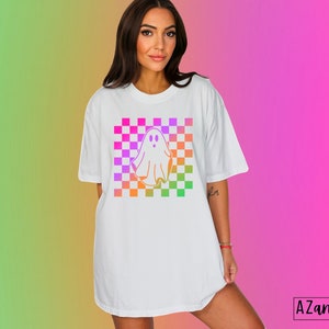 Op de afbeelding: Wit T-shirt met een kleurrijk ruitpatroon en een spookafbeelding. Het spookontwerp is omlijnd in een verloop van oranje, geel, roze en paars. Het shirt heeft korte mouwen en een relaxte pasvorm. De tekst "AZandAddi DESIGNS" staat rechtsonder.