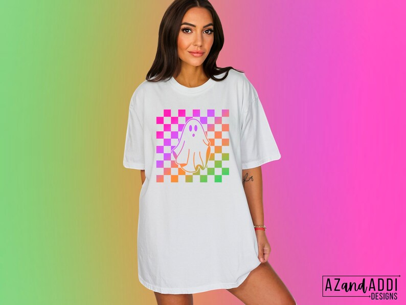 Op de afbeelding: Wit T-shirt met een kleurrijk ruitpatroon en een spookafbeelding. Het spookontwerp is omlijnd in een verloop van oranje, geel, roze en paars. Het shirt heeft korte mouwen en een relaxte pasvorm. De tekst "AZandAddi DESIGNS" staat rechtsonder.