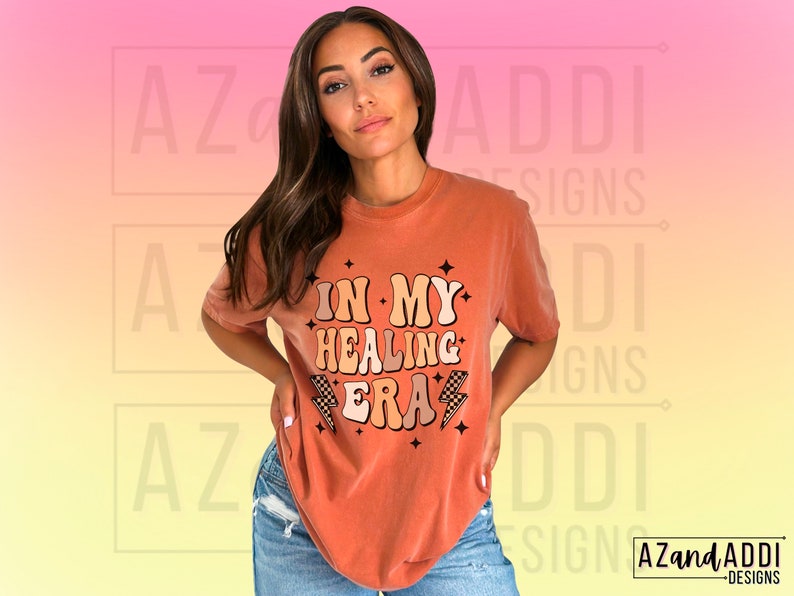 K&ouml;nnte beinhalten: Eine Frau tr&auml;gt ein orangefarbenes T-Shirt mit dem Text "In My Healing Era" in einer Retro-Schriftart mit einem Blitz-Motiv. Das Shirt ist locker geschnitten und hat einen Rundhalsausschnitt.