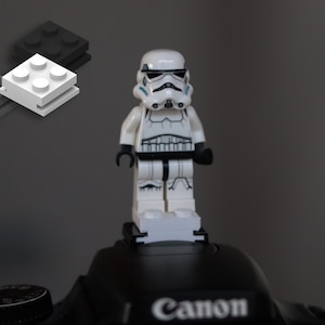 Op de afbeelding: Een witte Lego Stormtrooper-figuur met zwarte accenten en een gedetailleerde helm. De figuur staat op een witte Lego-steen, met grijze en zwarte stenen op de achtergrond. De figuur staat op een zwarte camera.