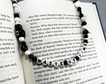 May include: A necklace made of black, white, and clear beads, featuring an 8-ball charm. The necklace spells "WASTE OF TIME" with white letter beads. It is displayed over an open book with printed text.