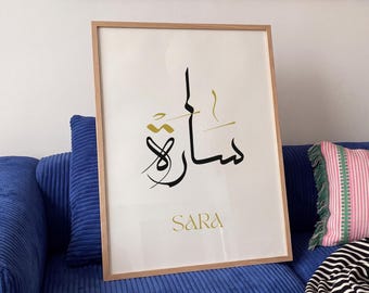 May include: A framed wall art print featuring black and gold Arabic calligraphy of the name Sara, with the name SARA printed in gold capital letters underneath. The artwork is encased in a light-colored wooden frame with a white background.