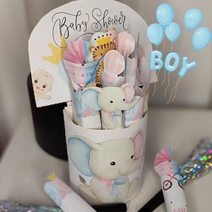 Könnte beinhalten: Ein Babyparty-Mittelstück mit einem weißen Behälter, der mit einer Elefantenillustration verziert ist. Der Behälter hat ein Pop-up-Design mit Baby-Themen-Ausschnitten und dem Wort "BOY" in blauen Ballonbuchstaben. Auf der Oberseite des Behälters steht "Baby Shower".