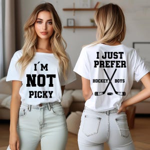 Könnte beinhalten: Ein weißes kurzärmeliges T-Shirt mit schwarzem Text auf der Vorderseite, der I'M NOT PICKY lautet. Die Rückseite des Shirts zeigt den Text I JUST PREFER HOCKEY BOYS über einer Grafik von zwei gekreuzten Hockeyschlägern und einem schwarzen Puck.