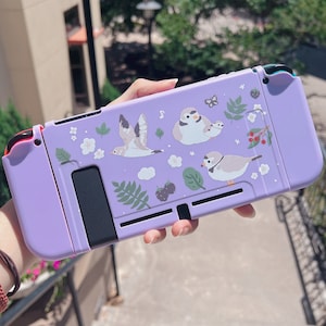 以下が含まれることがあります： 鳥と花の模様が施された、ラベンダー色のSwitchコンソールケース。ケースはマット仕上げで、保護設計です。デザインには、鳥、葉、花、ベリーのイラストが含まれています。ケースはSwitchコンソールを保護するように設計されています。