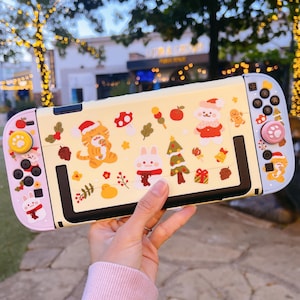 May include: A decorative yellow Nintendo Switch case featuring festive holiday-themed stickers. The case has a black frame and pink and blue accents. The stickers include a tiger, rabbit, and bear in Santa hats, a Christmas tree, and other holiday icons.