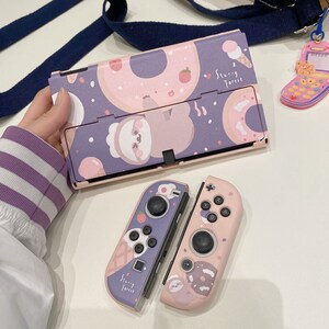 Op de afbeelding: Een roze en paarse Nintendo Switch-console en twee bijpassende Joy-Con-controllers met een luiaard- en donutontwerp. De console en controllers hebben de tekst "Starry Forest".