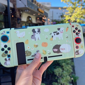 May include: A light green Nintendo Switch case with a farm animal theme. The case features a cow, sheep, duck, bee, and dog. The case is decorated with white flowers and green leaves.