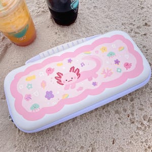 以下が含まれることがあります： 漫画のウーパールーパーと海の生き物が描かれた白とピンクのケース。ケースは薄紫色の縁取りと白いハンドルが付いています。ストローの入った透明なプラスチックカップに入った2つの飲み物が背景にあります。