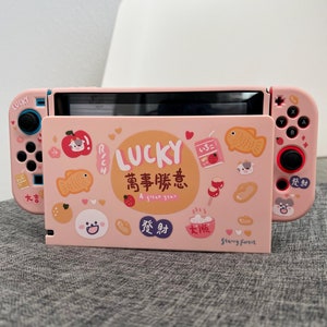 May include: Pink Nintendo Switch case with a Japanese-inspired design featuring a lucky cat, fish, and other good luck charms. The case has the text "LUCKY 萬事勝意 A great year" printed on it.
