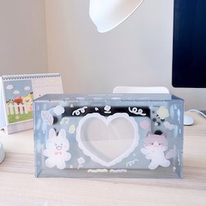 May include: A clear plastic tissue box with a heart-shaped opening and a cute cartoon bunny and cat design. The box has a white background with pastel pink, yellow, and blue accents.