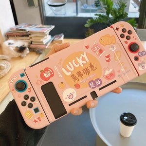 May include: A pink Nintendo Switch case with a Japanese design featuring a lucky cat, strawberries, and the text "LUCKY 萬事勝意 A great year 發財".