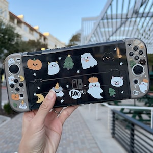 Peut inclure: Une coque transparente pour Nintendo Switch sur le thème d'Halloween. La coque est décorée de divers autocollants représentant des fantômes, des citrouilles et d'autres personnages sur le thème d'Halloween. Le texte "BOO!" est visible sur la coque.