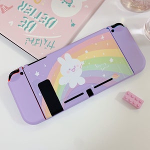 Peut inclure: Une coque Nintendo Switch de couleur violette et blanche avec un arc-en-ciel et un lapin blanc tenant une baguette magique. La coque porte l'inscription "Starry Turtle".