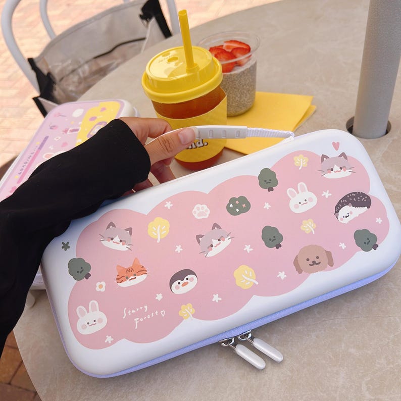 May include: A white and pink case with a cartoon design featuring cats, a bunny, a hedgehog, a penguin, and trees. The case has a light blue zipper and the text "Starry Forest".