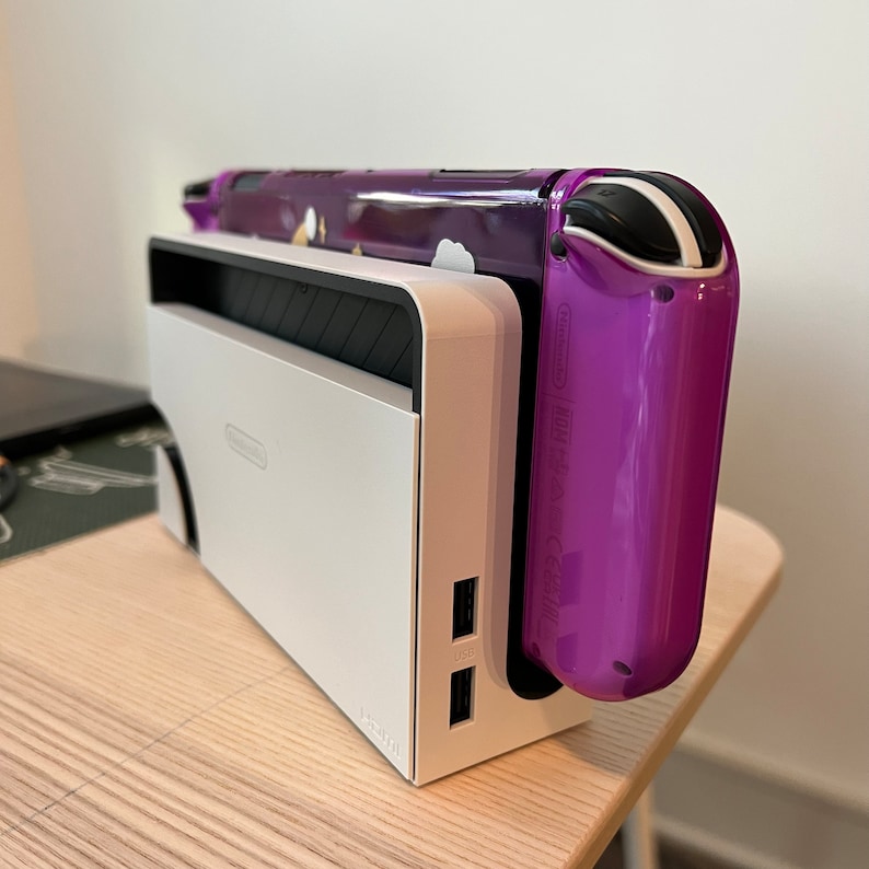 May include: A white Nintendo Switch charging dock with two USB ports. A purple Nintendo Switch with a clear case is in the dock.