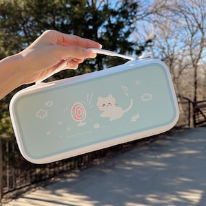 May include: A light blue rectangular case with a white handle and zip. The case features a cartoon cat design with a fan, fish, and the text "A very happy day".