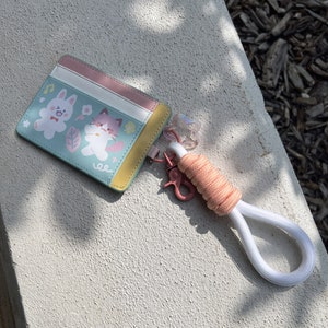 May include: A light blue card holder with a pink, white, and yellow trim. The card holder features a cartoon illustration of a bunny and a cat. The card holder is attached to a white and pink lanyard.
