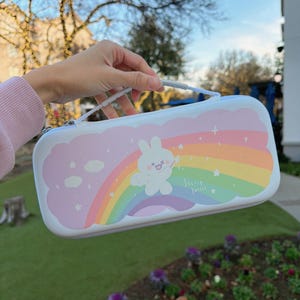 Pode incluir: Uma mala de transporte branca e rosa com um coelho de desenho animado, um arco-íris e o texto "Starry Forest". A mala tem uma alça e um fecho de correr. O design inclui cores pastel.