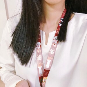 Może przedstawiać: Biała koszula z długim rękawem i dekoltem w kształcie litery V oraz brązowo-biały sznurek z kresk&oacute;wkowym psem i wzorem kawiarni. Sznurek ma nadrukowany tekst "La Vrise Cafe".